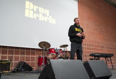 Eksmisbrugeren Julius står på scenen og taler. På storskærmen bag ham står der 'Drug Rebels' med gul skrift.