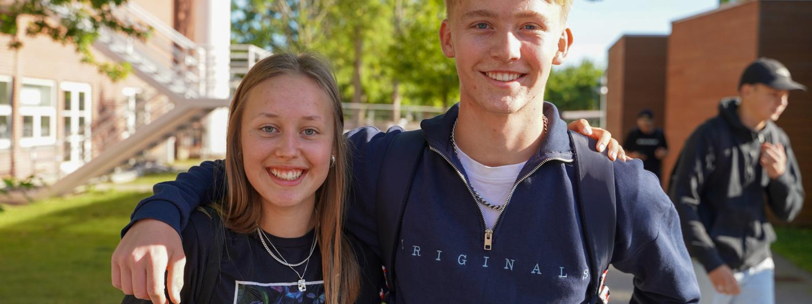 En dreng og en pige smiler til kameraet, men de holder om hinanden uden for skolen.