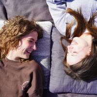 To piger ligger på et tæppe i solen og griner til hinanden.
