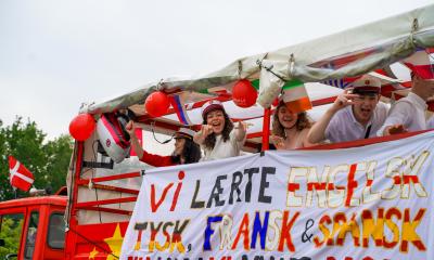 Glade studenter jubler mod kameraet i deres røde studentervogn med banner og balloner.