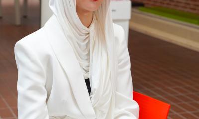 Portræt af en muslimsk studenterpige med hvidt tørklæde på under huen. Hun sidder  i kantinen og smiler til kameraet.